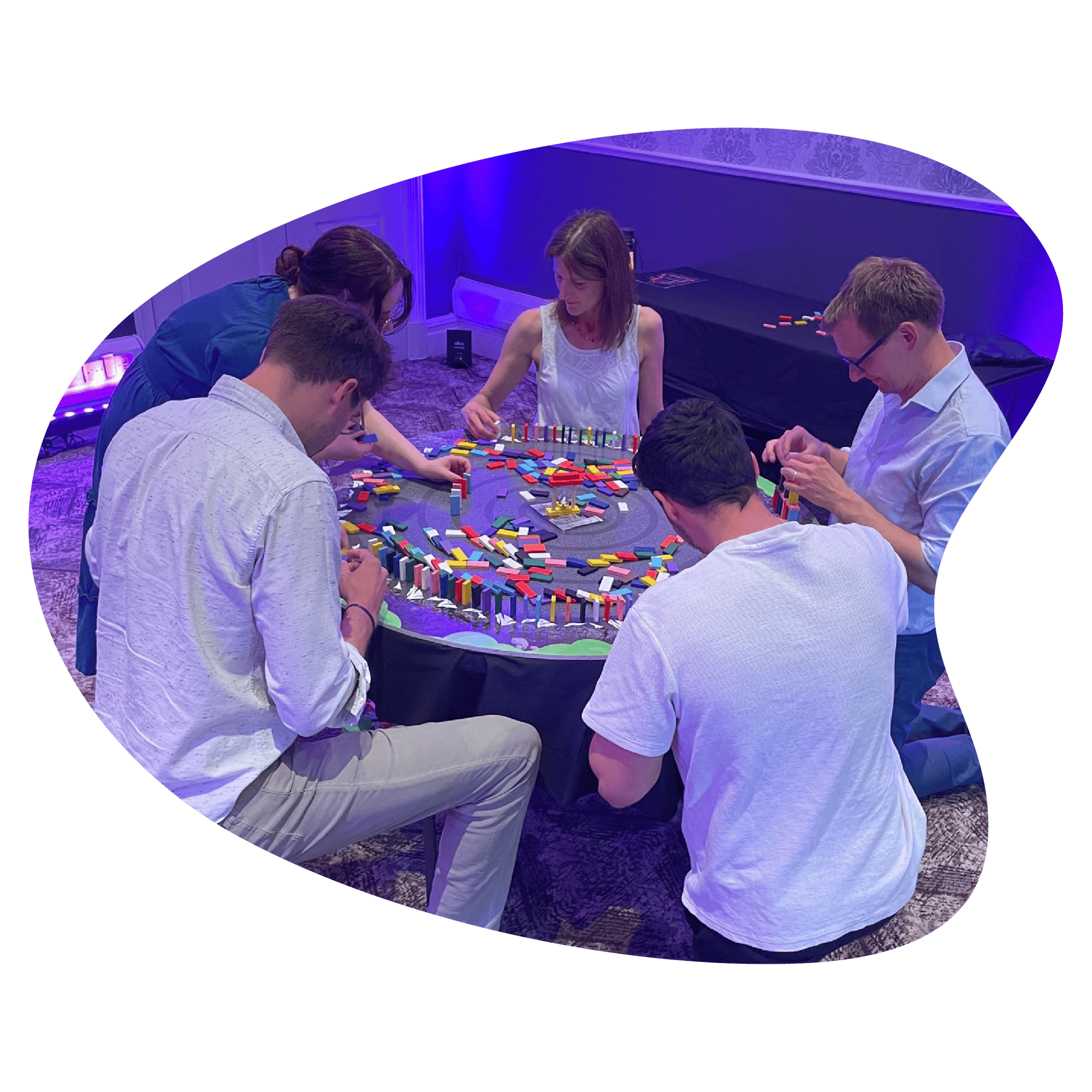 A group of five people are gathered around a table, stacking dominoes.