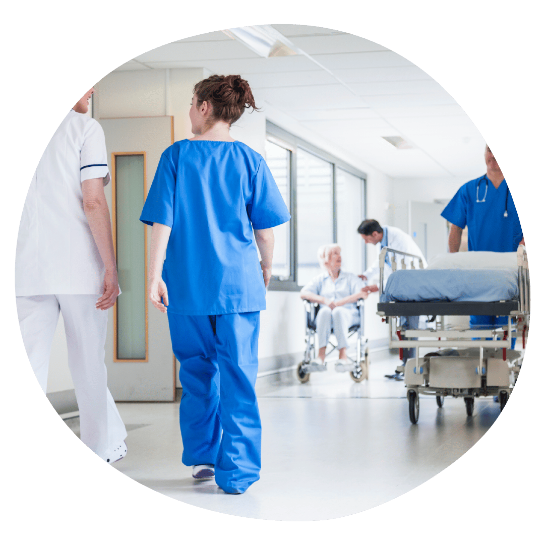 Clinicians are walking through a corridor. In the background is a person in a wheelchair and another clinician pushing an empty bed