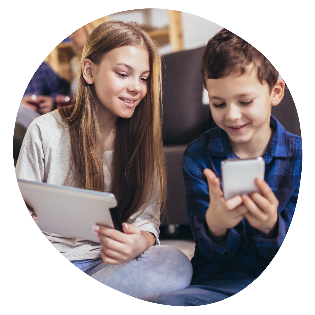 Two children are sat next to each other smiling. One is using a tablet, while the other is looking at a phone