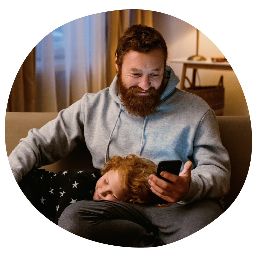 A child is laid in their parent's lap on the sofa. The parent is smiling looking at their phone A child is laid in their parent's lap on the sofa. The parent is smiling looking at their phone