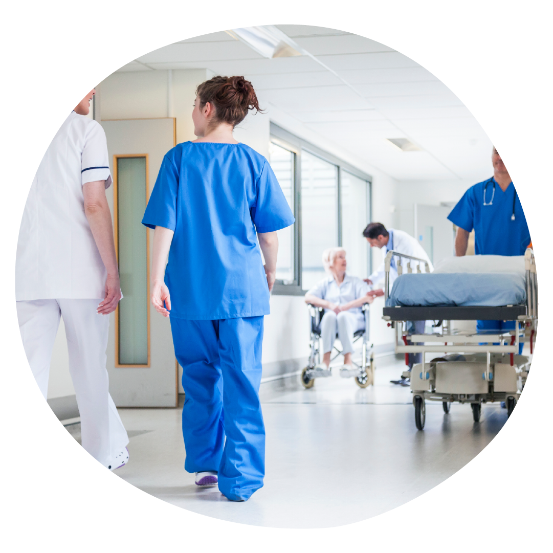 Clinicians are walking through a corridor. In the background is a person in a wheelchair and another clinician pushing an empty bed